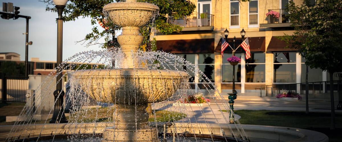 A multi-tiered stone fountain with water jets, surrounded by a paved area with a building in the background.