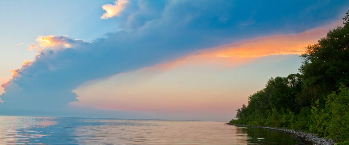 A calm lake at sunset with a rocky shoreline and dense trees on the right.