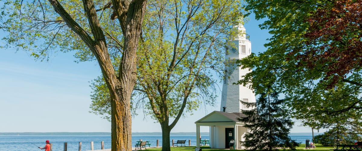 A park with a lighthouse, a bench, and a person fishing by the water.