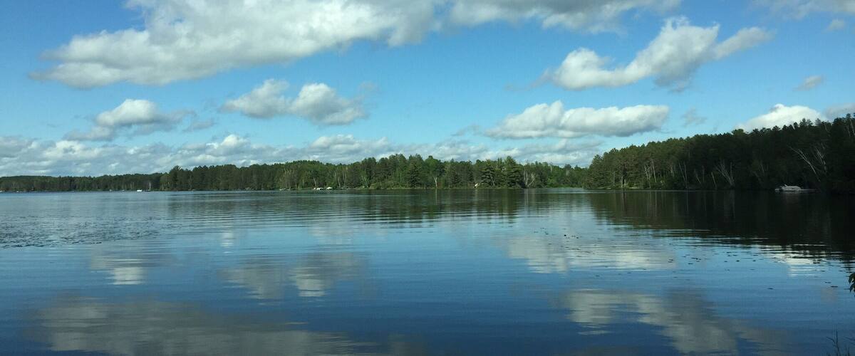 A calm lake with a clear reflection of the sky and clouds, surrounded by dense forest.