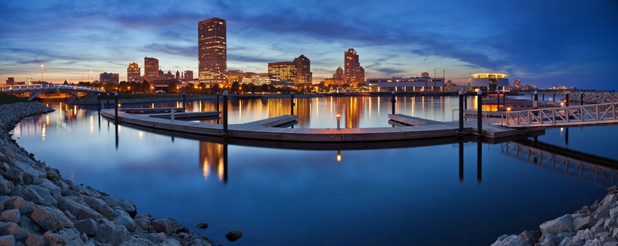 A waterfront cityscape at dusk with a calm river reflecting buildings and a bridge.