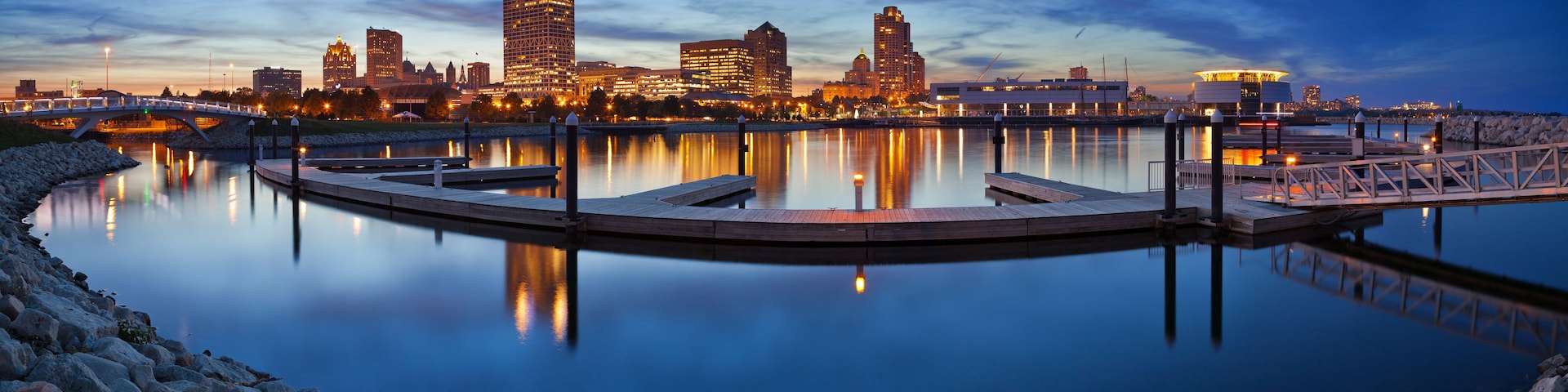 Paisaje urbano frente al río al atardecer, con un río tranquilo que refleja edificios y un puente.