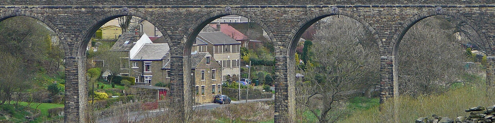 Un puente de piedra con tres arcos que cruza un río, rodeado de vegetación y edificios residenciales al fondo.