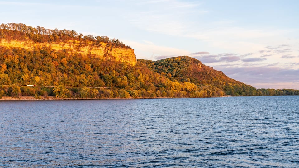 A river with a forested cliff and a small building on the shore.