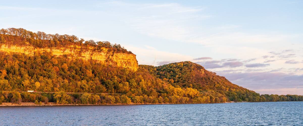A river with a forested cliff and a small building on the shore.