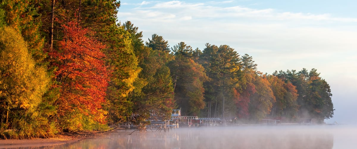 A calm lake with autumn-colored trees reflecting on the water, a misty shoreline, and a clear sky.
