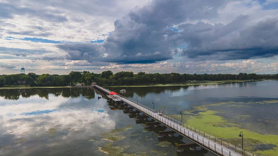 A long bridge over a calm lake with a red-roofed pavilion, surrounded by trees and a cloudy sky.