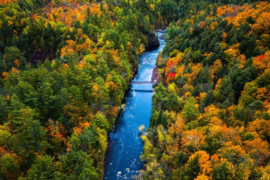 Beautiful travel aerial of a pedestrian foot bridge crossing the bright blue water of the Bad River at Copper Falls with colorful fall foliage lining the river banks in autumn in Mellen, Wisconsin.