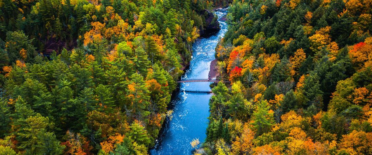 Beautiful travel aerial of a pedestrian foot bridge crossing the bright blue water of the Bad River at Copper Falls with colorful fall foliage lining the river banks in autumn in Mellen, Wisconsin.