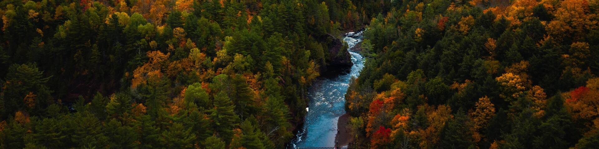 Beautiful aerial above the Bad River and a pedestrian foot bridge at Copper Falls with colorful fall foliage lining the river banks and cloudy sky above the horizon in autumn in Mellen, Wisconsin.