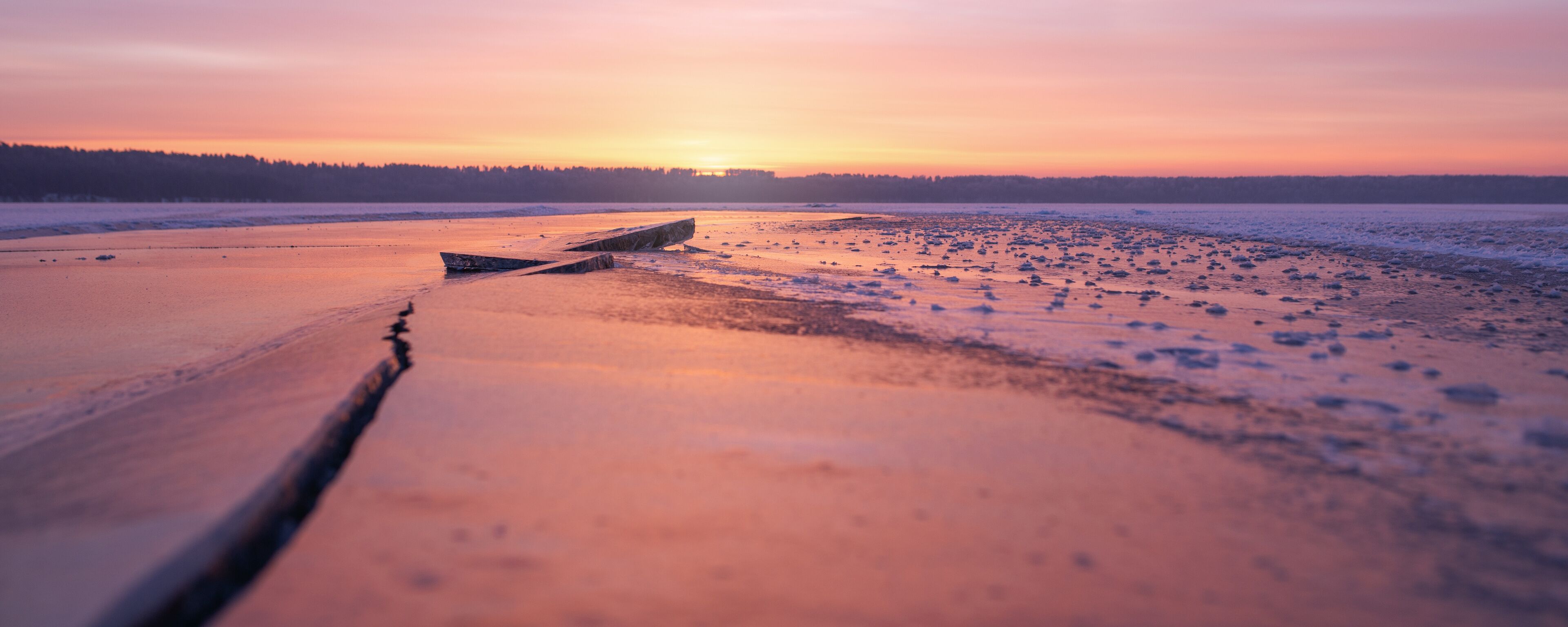 A frozen lake at sunset with a wooden boat and a forest in the background.