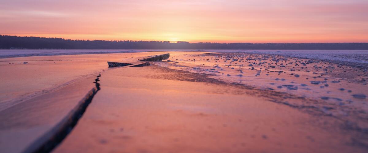 A frozen lake at sunset with a wooden boat and a forest in the background.