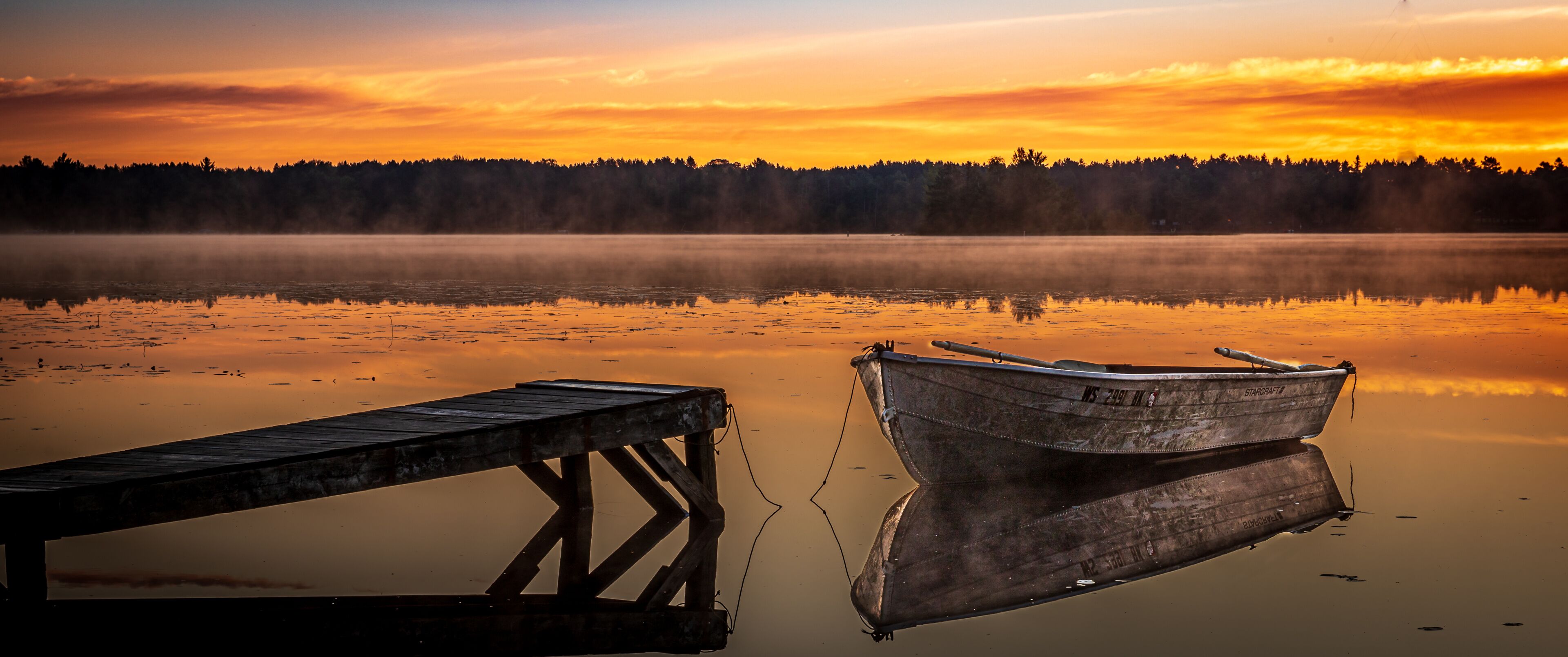 A wooden dock and a rowboat on a calm lake at sunrise with a forest in the background.