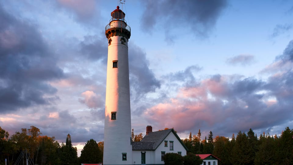 A white lighthouse with a red top, a small house with a green door, and a grassy field with trees in the background.