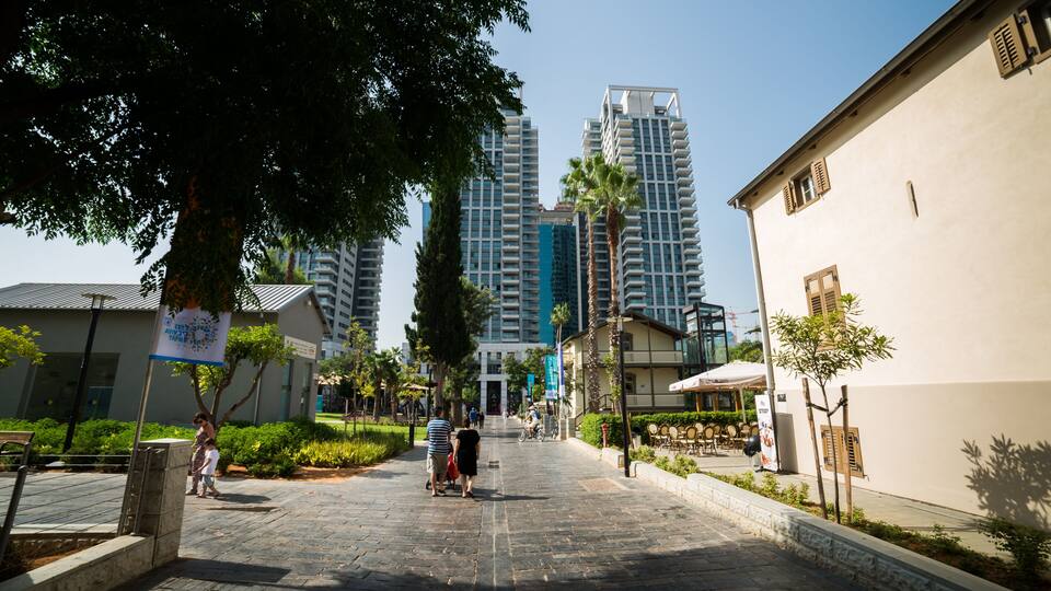 A paved walkway lined with trees and buildings, including a tall modern structure in the background.