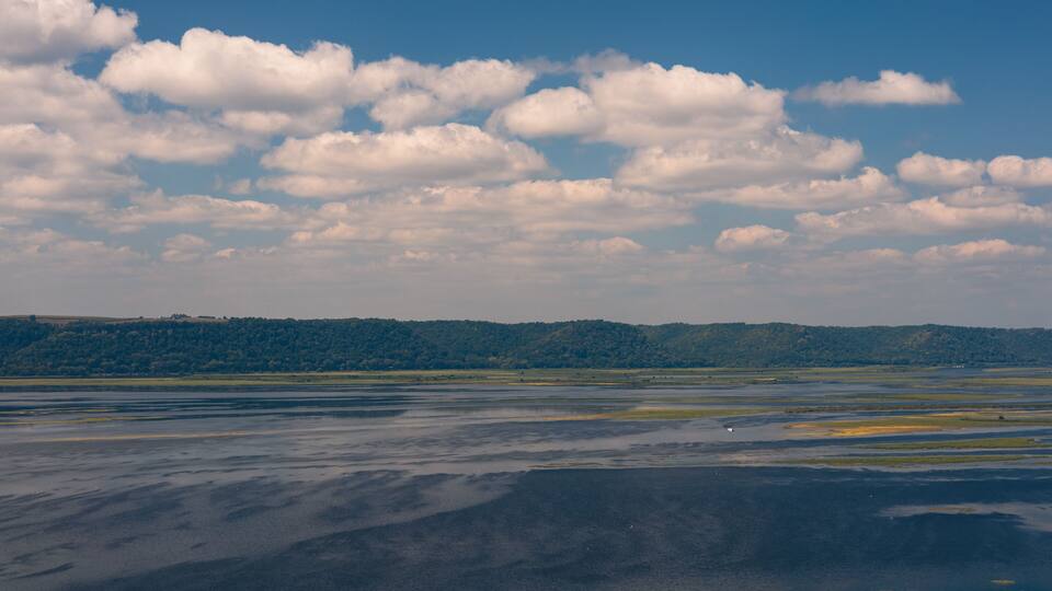 A landscape with a body of water reflecting the sky, a distant forested hill, and scattered clouds.