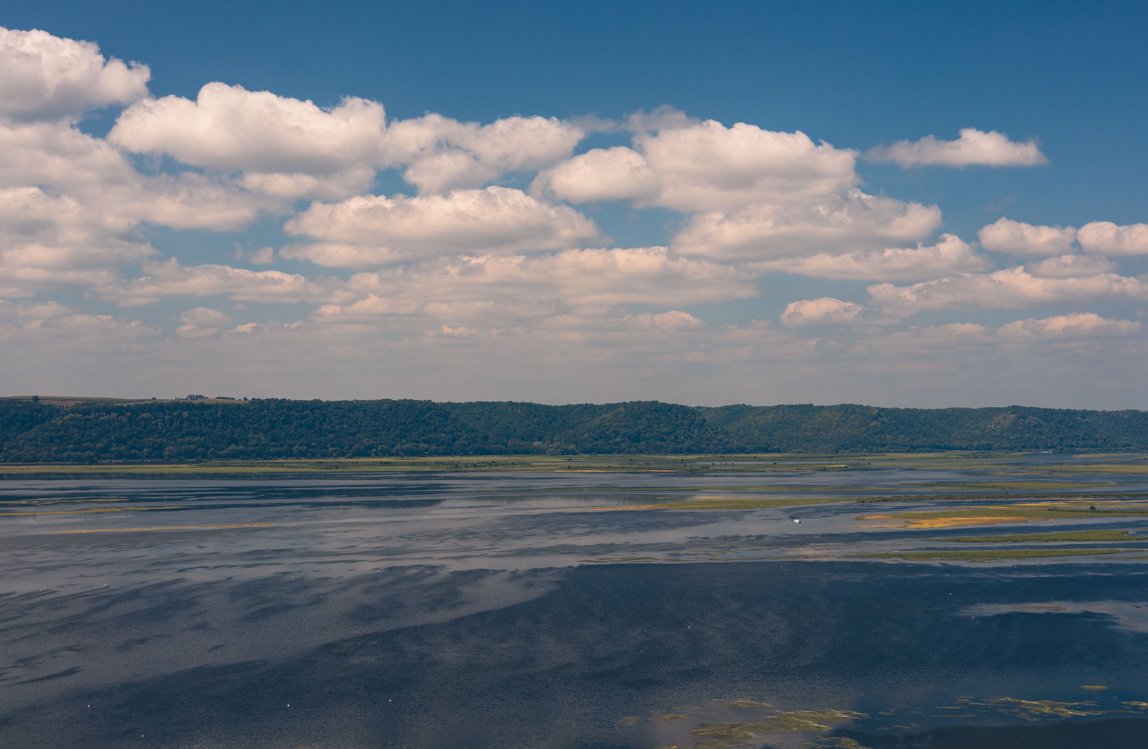 A landscape with a body of water reflecting the sky, a distant forested hill, and scattered clouds.