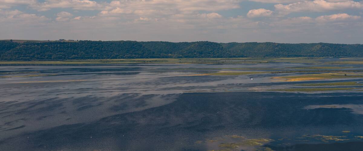 A landscape with a body of water reflecting the sky, a distant forested hill, and scattered clouds.
