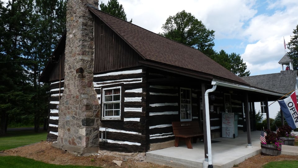 A log cabin with a stone chimney, surrounded by greenery and a clear sky.