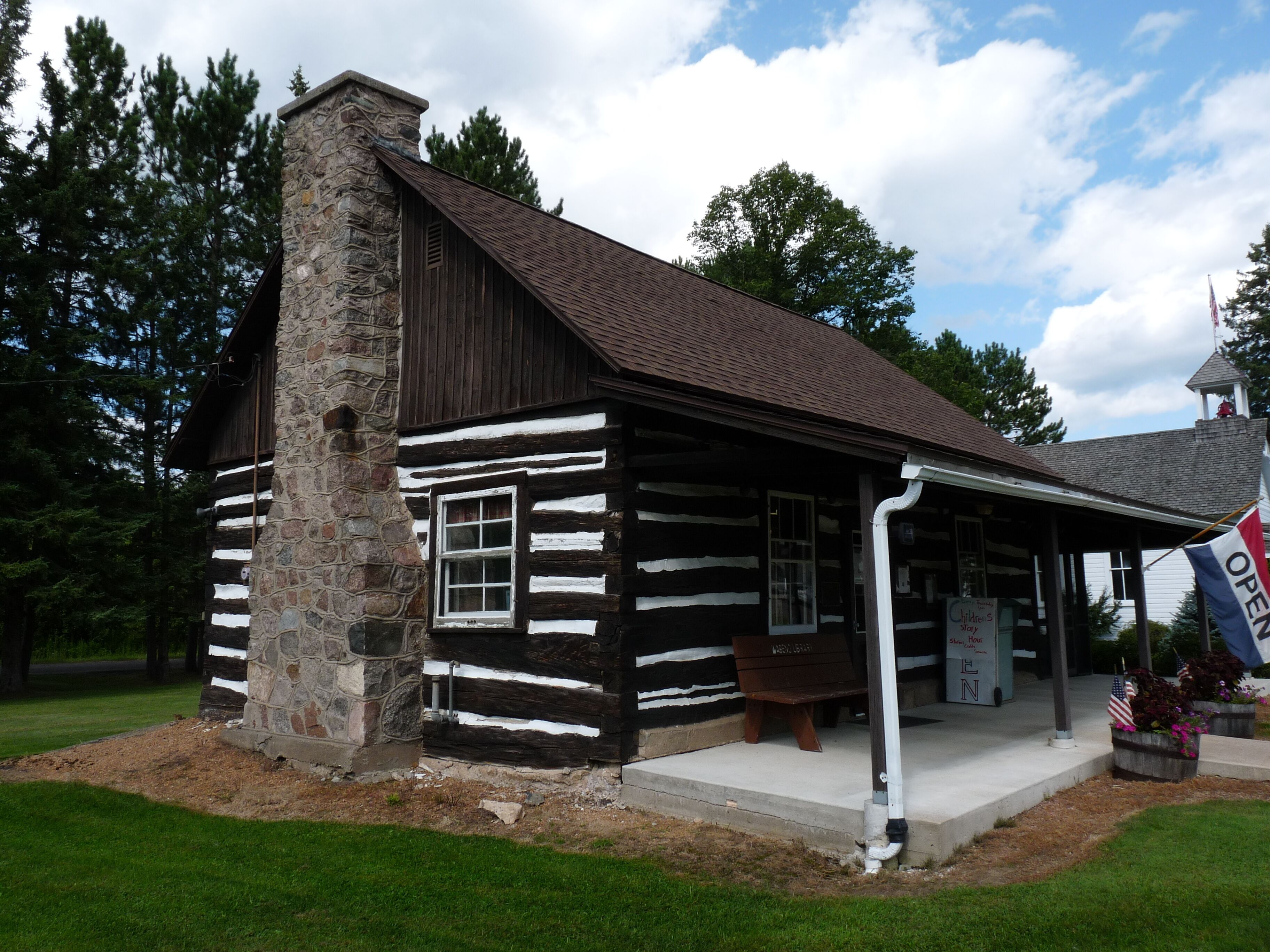 A log cabin with a stone chimney, surrounded by greenery and a clear sky.