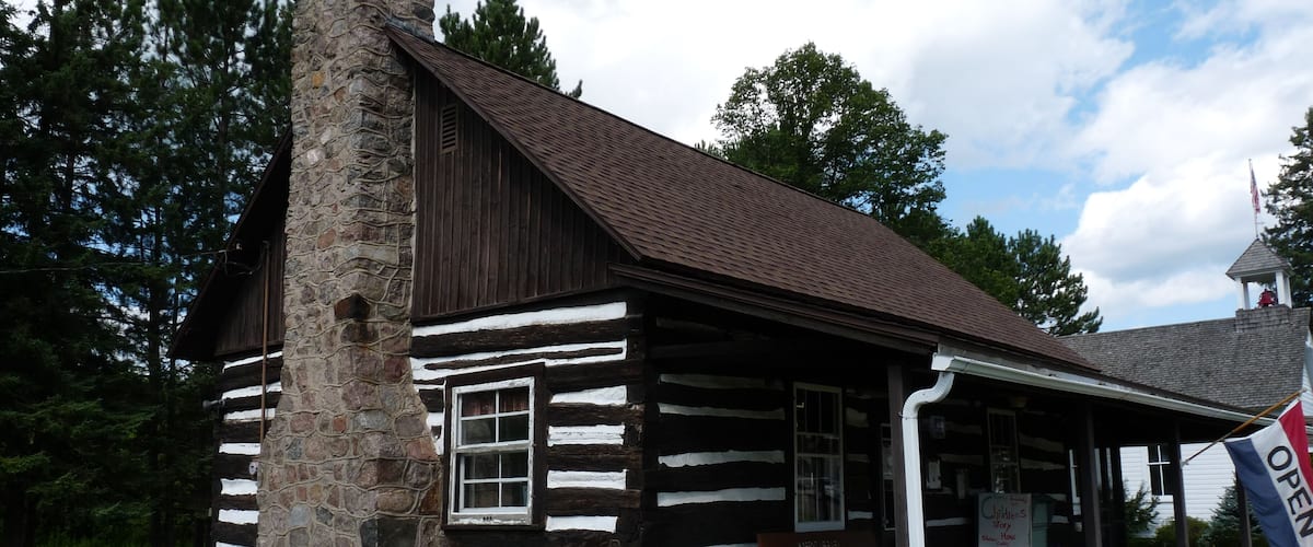 A log cabin with a stone chimney, surrounded by greenery and a clear sky.
