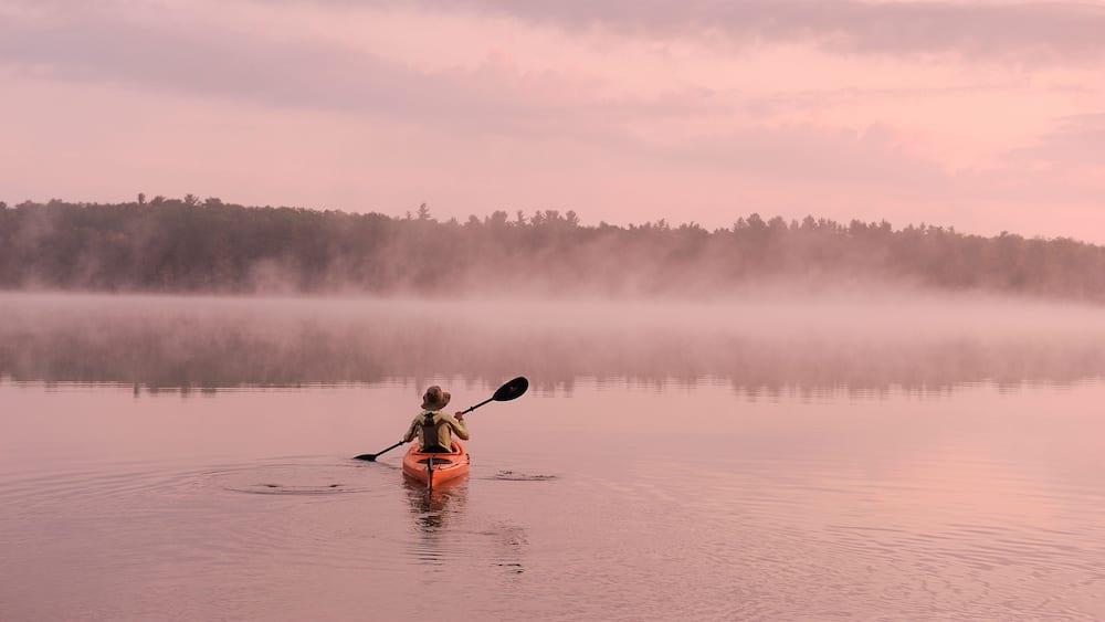A kayaker in a calm body of water with a forested shoreline and a misty sky.