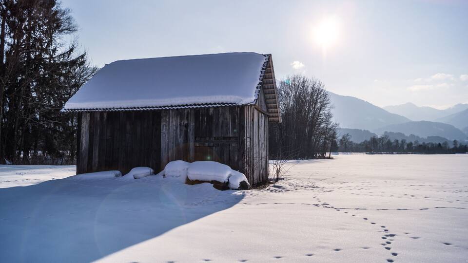 A wooden cabin with snow-covered roof and steps, surrounded by a snowy landscape and mountains in the background.