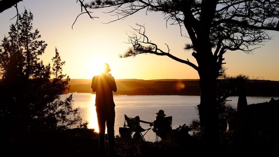 A silhouette of a person standing and sitting on a lakeside at sunset.