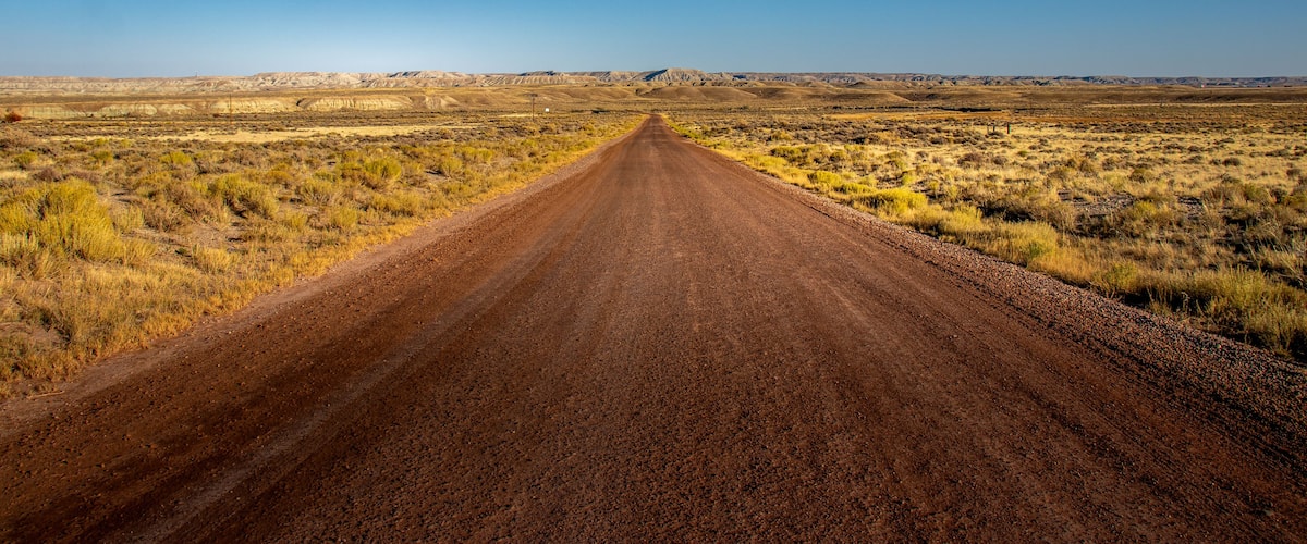 A long, straight dirt road in a flat, open landscape with sparse vegetation and distant hills under a clear blue sky.