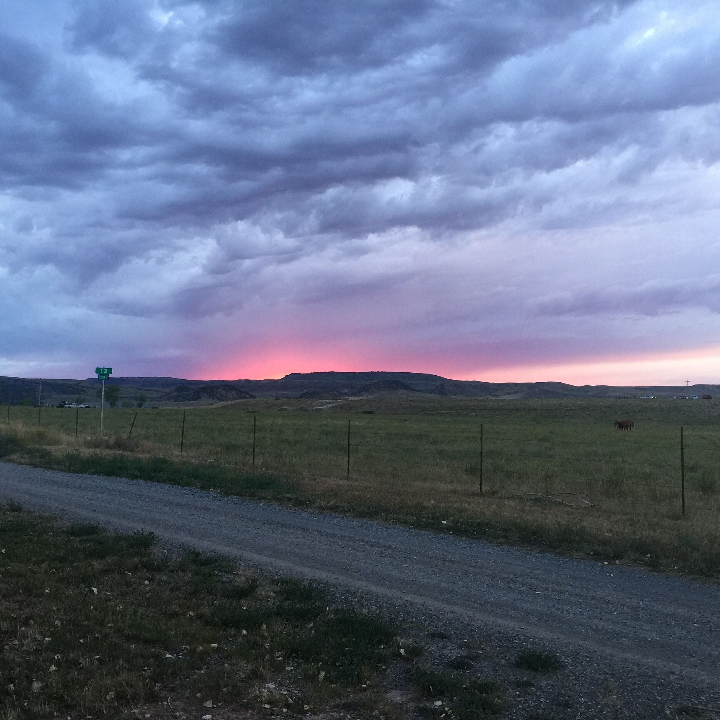 A rural landscape with a dirt road, a green signpost, and a sunset over rolling hills.