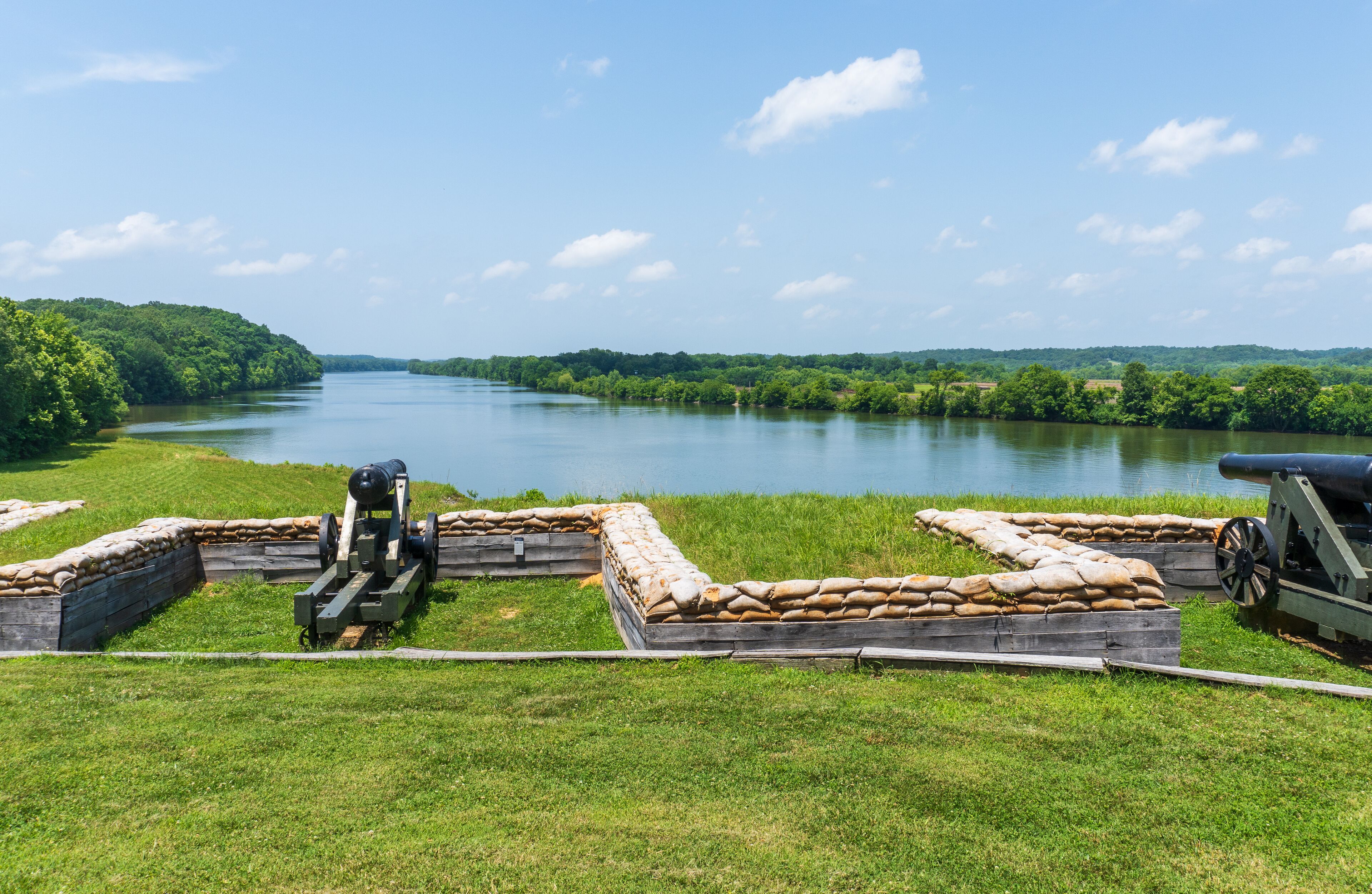 A cannon on a grassy field overlooking a calm river with a forested riverbank.