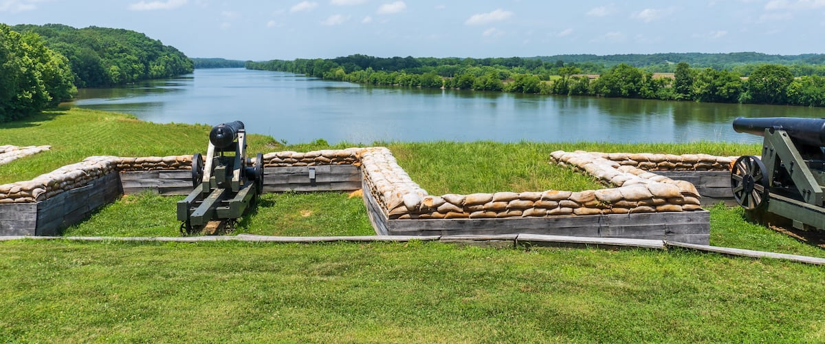 A cannon on a grassy field overlooking a calm river with a forested riverbank.