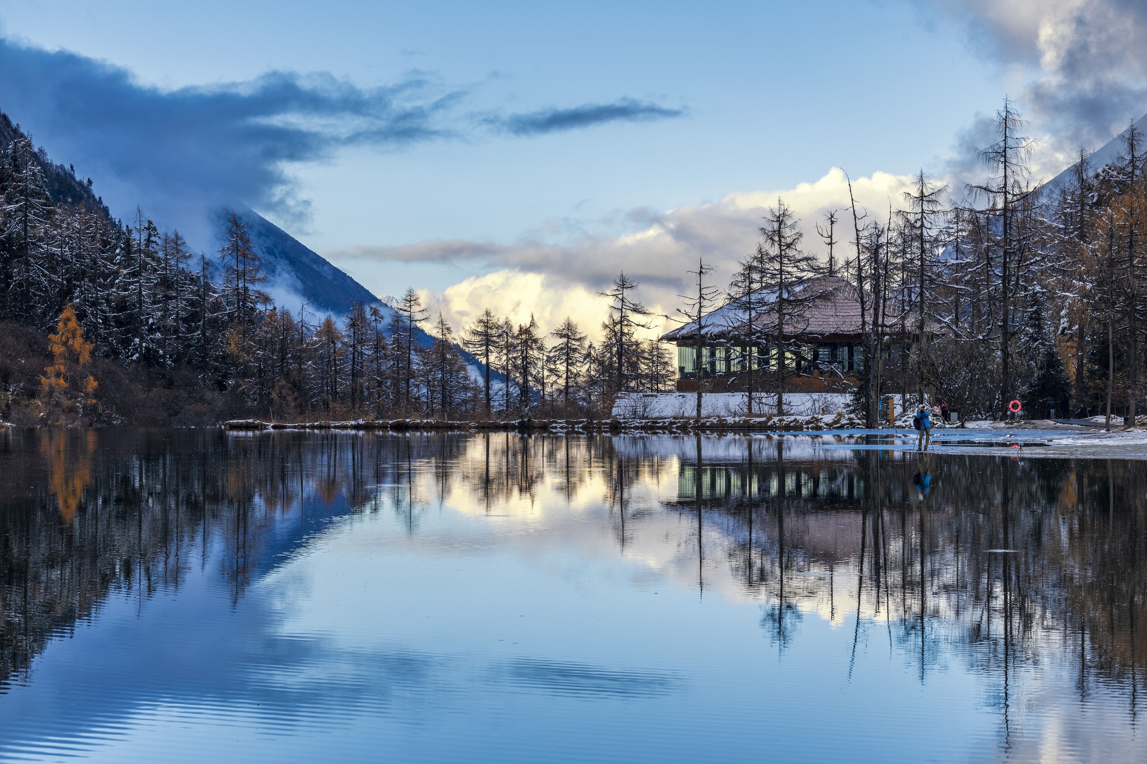 Ein ruhiger See mit Spiegelung von Bäumen und einer Bergkette, eine Reetdachhütte und ein klarer Himmel.