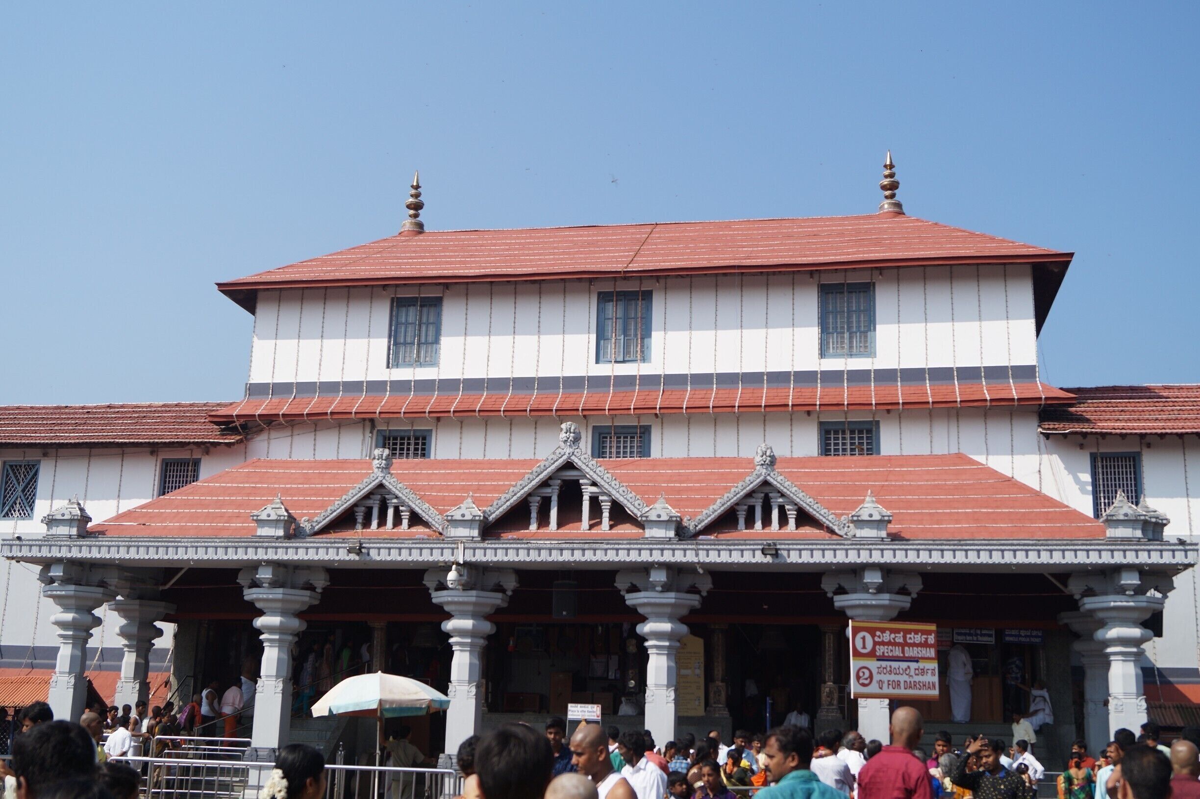 A large, traditional building with a red roof and white walls, featuring ornate architectural details and a crowd of people in front.
