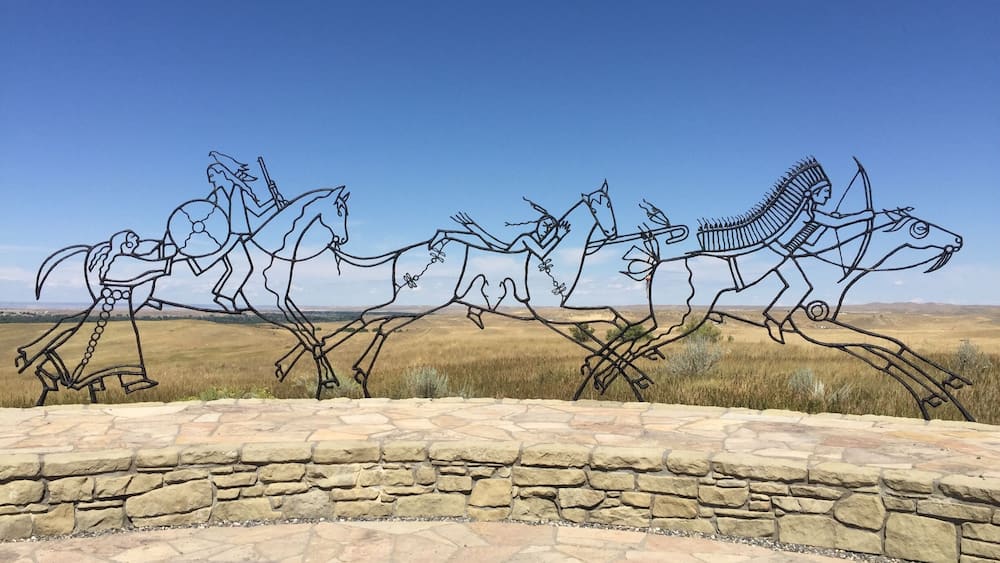 A metal sculpture of three horses in motion, set against a clear blue sky and a flat landscape.