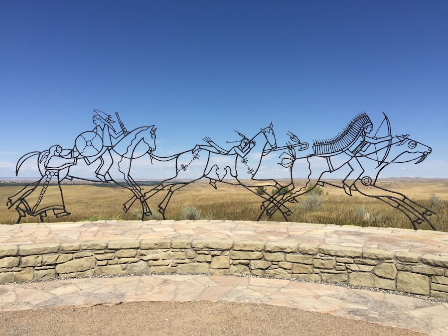 A metal sculpture of three horses in motion, set against a clear blue sky and a flat landscape.
