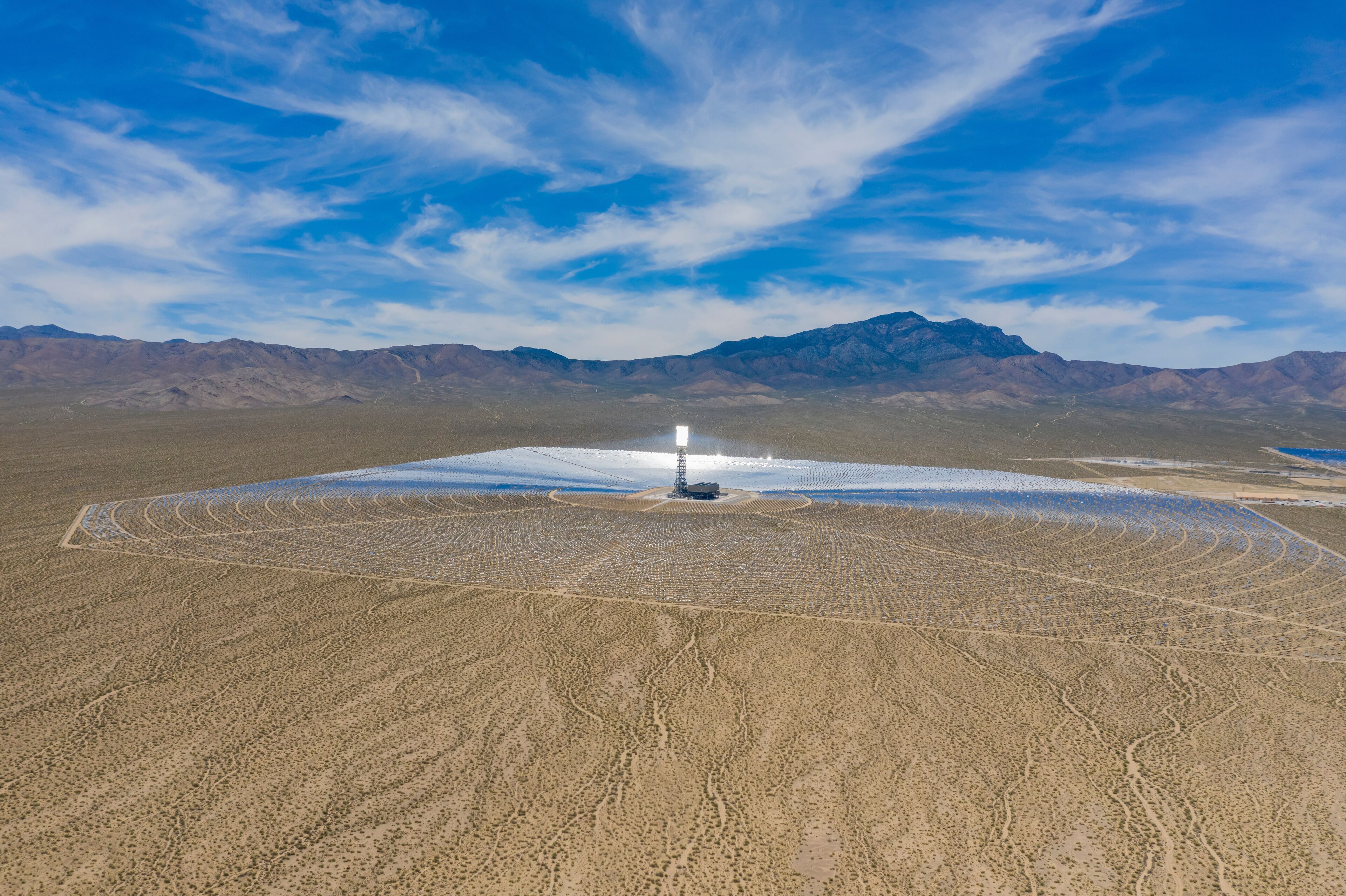 A vast desert landscape with a circular pattern of disturbed ground, a central structure, and distant mountains under a clear sky.