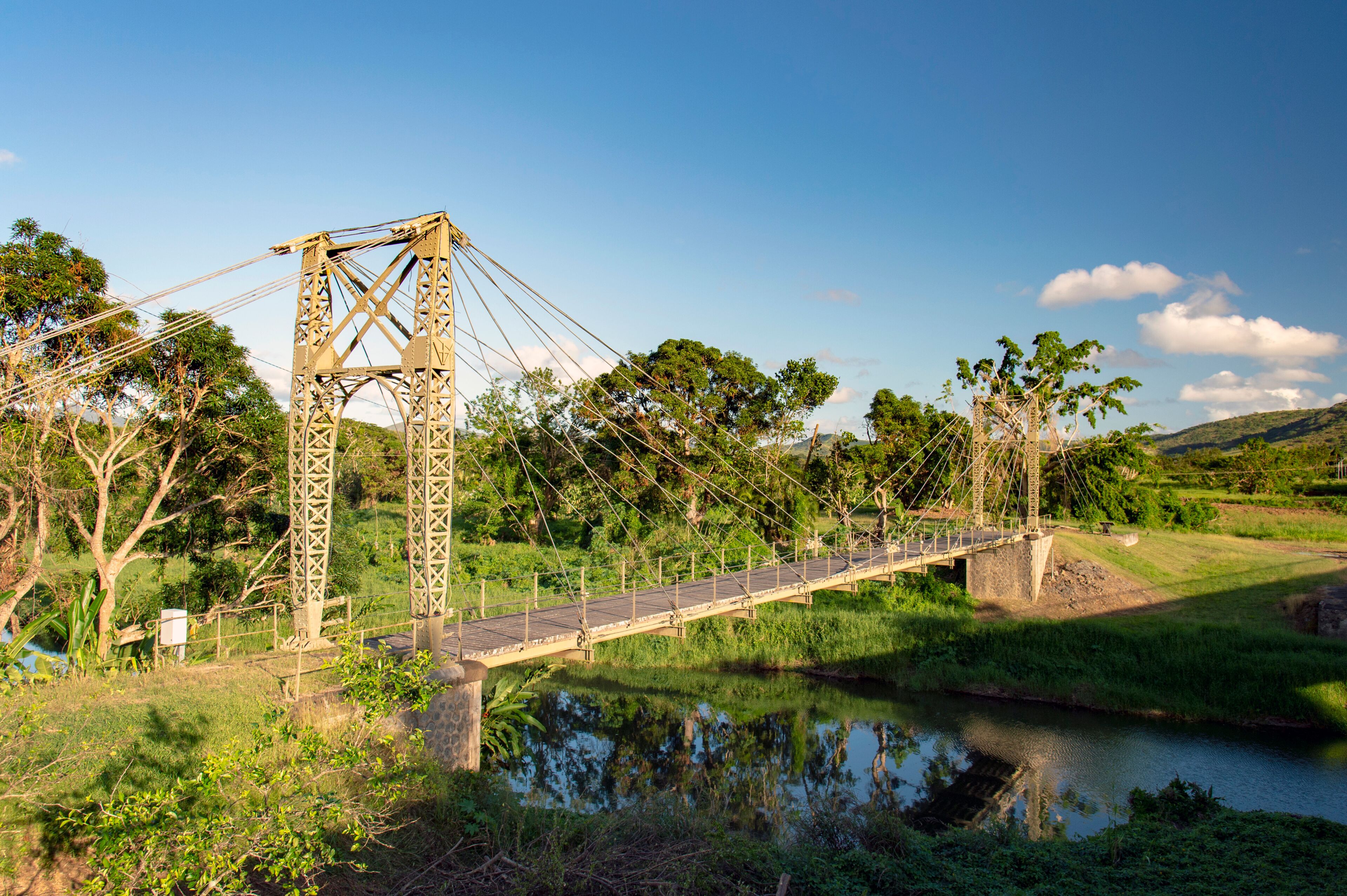 A suspension bridge over a calm river, surrounded by lush greenery and trees.