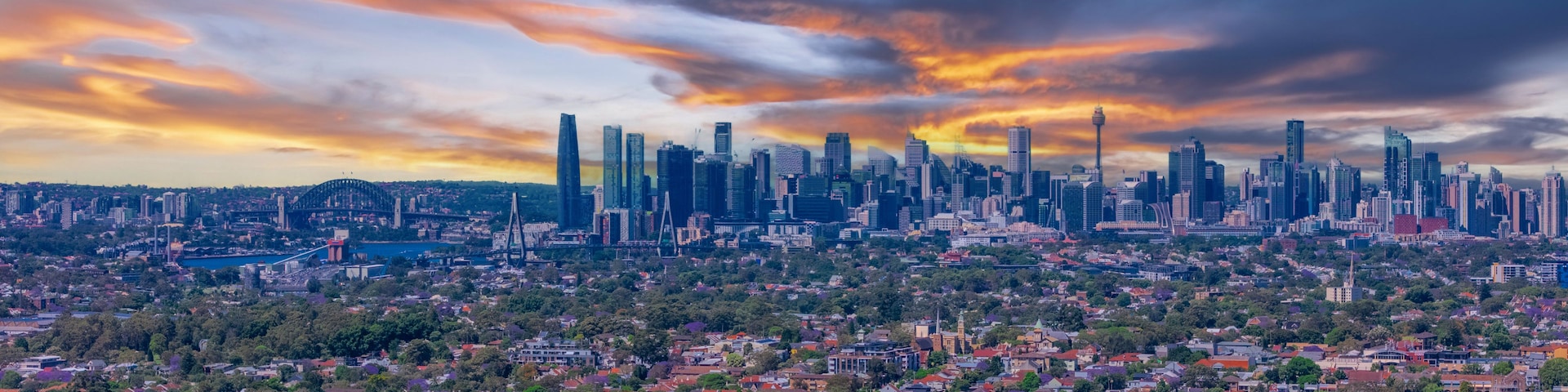 A cityscape with a dense urban area, a prominent bridge, and a skyline with high-rise buildings under a sunset sky.