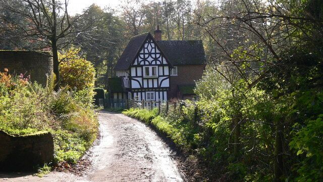 A narrow rural road leading to a Tudor-style house surrounded by lush greenery.