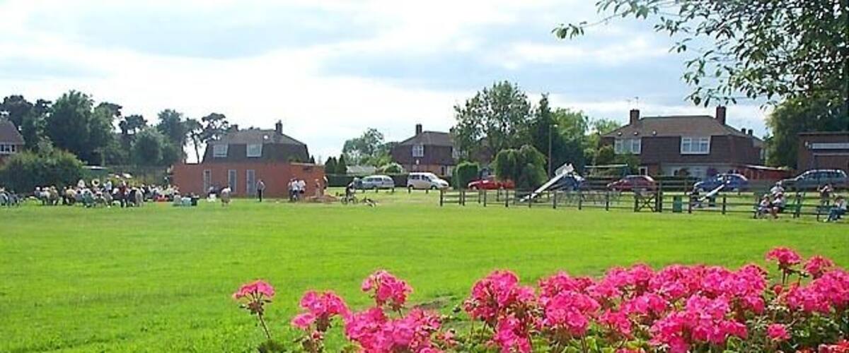 A grassy field with a flower bed in the foreground and houses in the background.