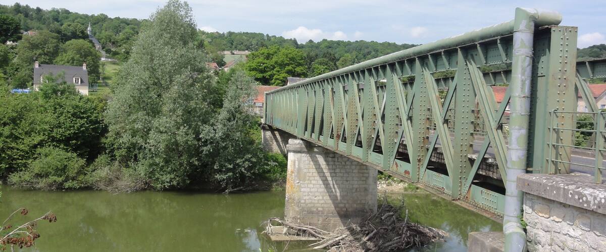 A green metal bridge over a river with trees and houses in the background.