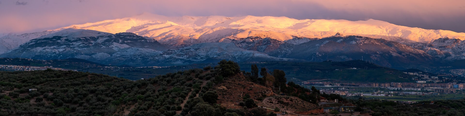 A snow-capped mountain range at sunset with a town in the valley below.