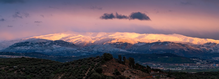 A snow-capped mountain range at sunset with a town in the valley below.