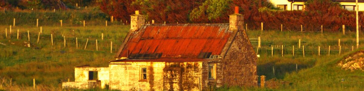 Ein rustikales Steinhaus mit rotem Dach in einer grünen Wiese, umgeben von Bäumen und einem Berg im Hintergrund.