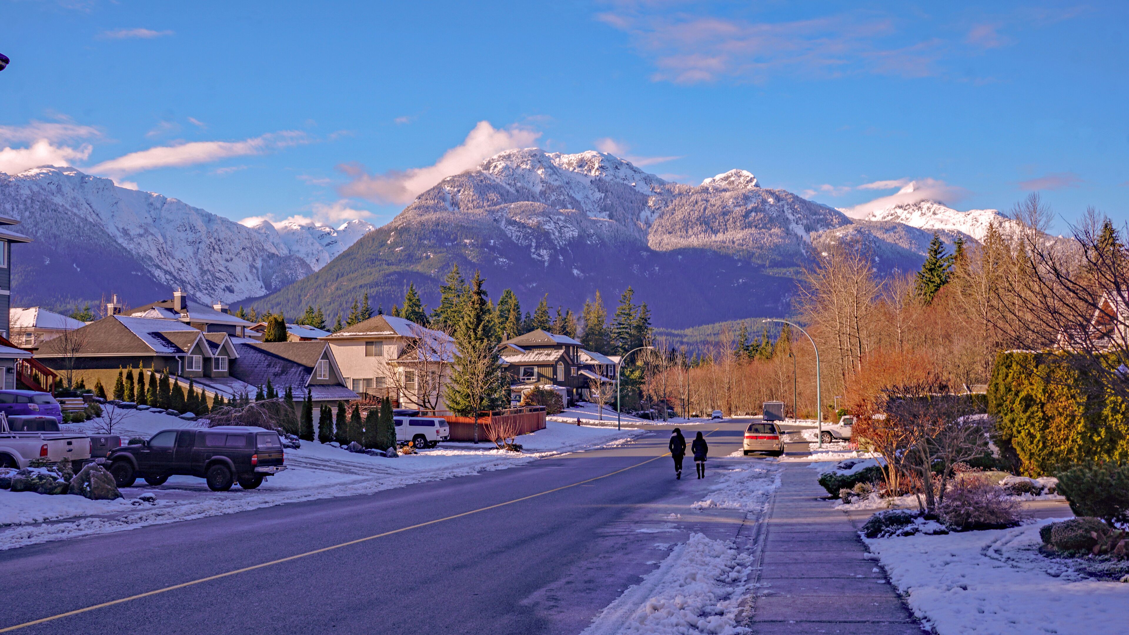 A snow-covered street in a residential area with houses, parked cars, and mountains in the background.