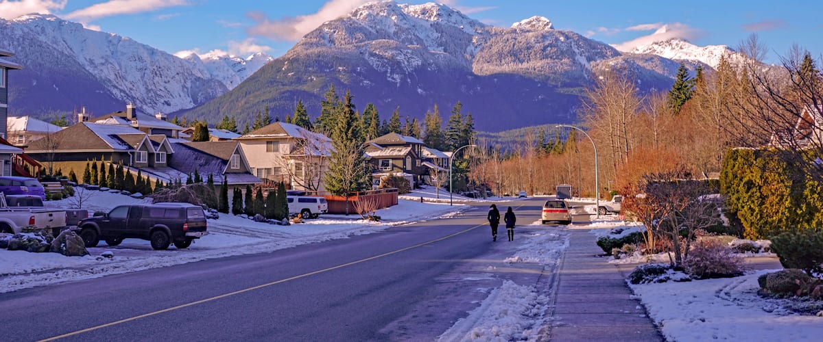 A snow-covered street in a residential area with houses, parked cars, and mountains in the background.