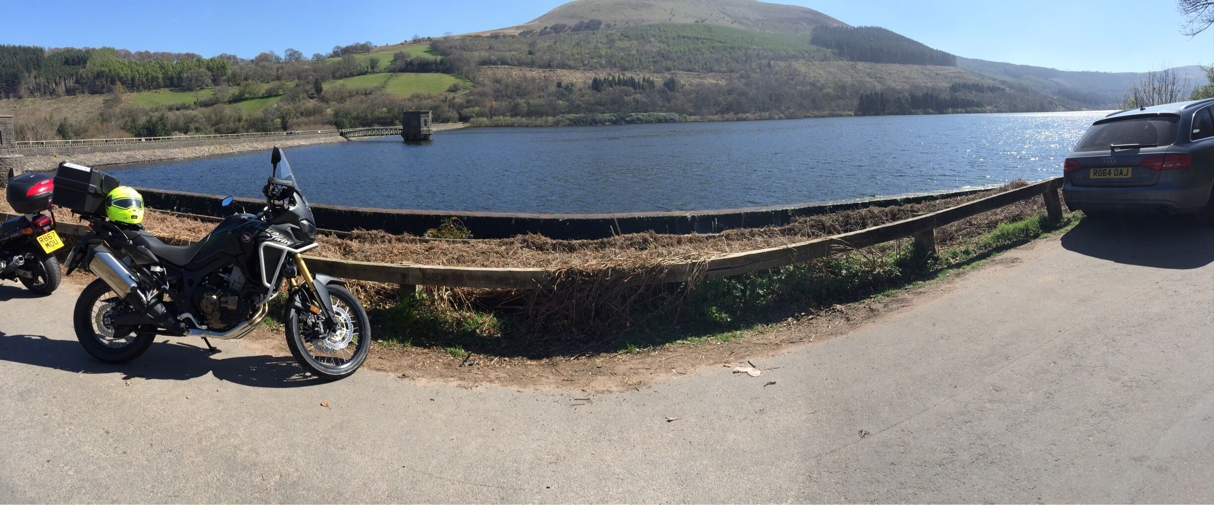 A motorcycle parked by a lake with a wooden fence and a car in the background.