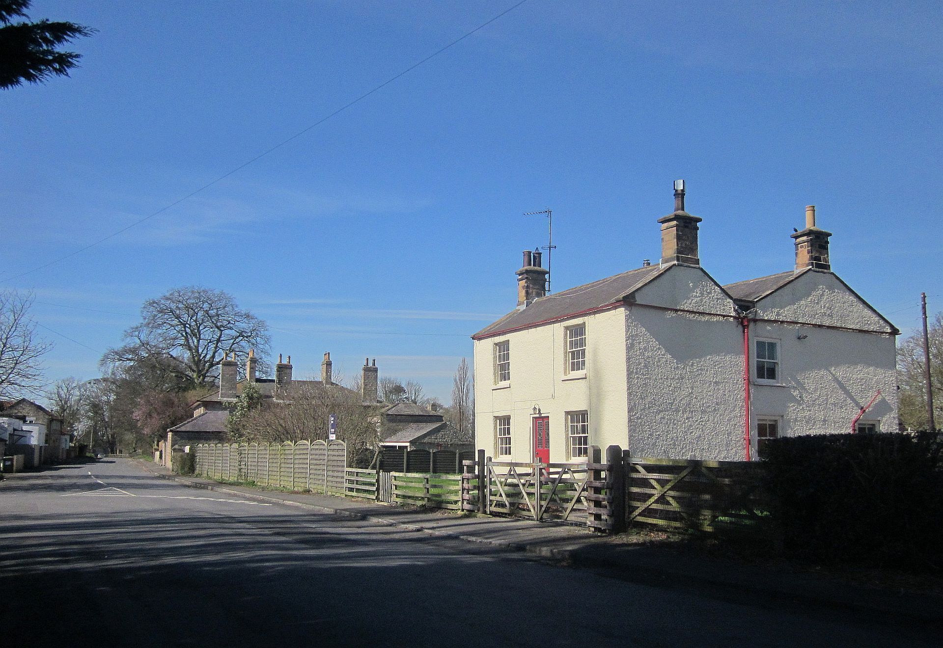 A rural street with a white house with a red door, a wooden fence, and a clear blue sky.