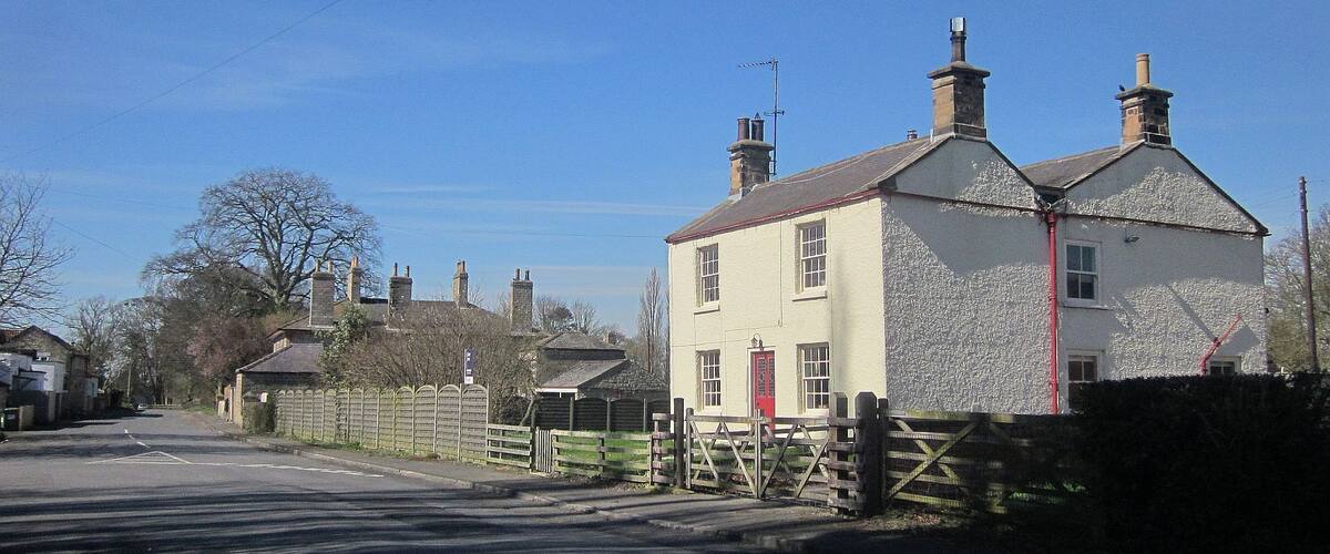 A rural street with a white house with a red door, a wooden fence, and a clear blue sky.
